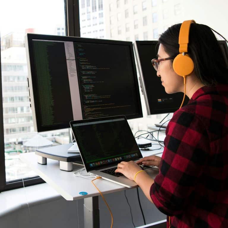 woman with headphones on at at workstation with two monitors and a laptop. Coding is on the screen.